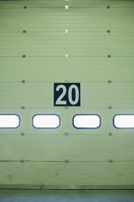 A large green industrial garage door with horizontal paneling, featuring a white and black number '20' sign prominently displayed in the center. Below the number, there are four oval-shaped windows wi