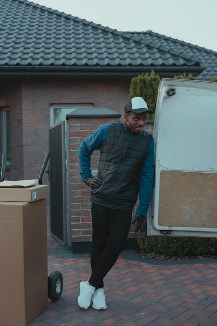 A man wearing a dark vest, blue long-sleeve shirt, black pants, black gloves, white sneakers, and a grey cap stands outside a brick house with a tiled roof, near a white moving van parked on a paved driveway. He leans against the van's open side door, which has a partially visible cardboard panel, during a home relocation or furniture transport process. In the foreground, there are large cardboard boxes, one on a wheeled dolly, likely packed for moving. The scene is set in natural daylight, and the background includes a small garden with shrubs and a neighbouring property, illustrating the loading or packing stage of a house removal. This image, associated with [COMPANY_NAME], showcases professional packing and moving activities typical of removals services near Edgware station, aligning with the theme of packing and loading for home relocation.