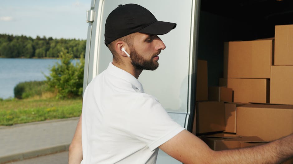 A man wearing a black cap, white T-shirt, and wireless earbuds is seen loading cardboard boxes into the back of a white moving van parked outdoors near a body of water and green trees. The van's open door reveals neatly stacked boxes of varying sizes, some wrapped in protective packing materials, indicative of a home relocation or furniture transport process. The man is captured in profile, focusing on carefully placing the boxes, with the surrounding environment illuminated by daylight, suggesting a clear, bright day suitable for packing and moving activities. This scene illustrates professional house removal services provided by Man with Van Edgware, emphasizing the logistics involved in packing, loading, and transporting household items in a systematic manner for a smooth move.