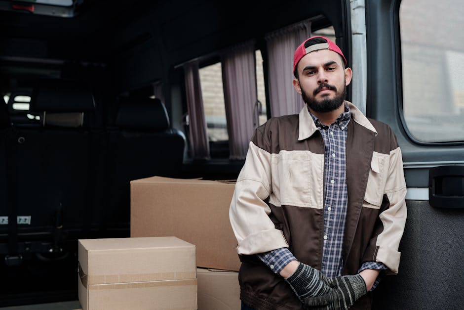 A man wearing a dark vest, blue long-sleeve shirt, black pants, black gloves, white sneakers, and a grey cap stands outside a brick house with a tiled roof, near a white moving van parked on a paved driveway. He leans against the van's open side door, which has a partially visible cardboard panel, during a home relocation or furniture transport process. In the foreground, there are large cardboard boxes, one on a wheeled dolly, likely packed for moving. The scene is set in natural daylight, and the background includes a small garden with shrubs and a neighbouring property, illustrating the loading or packing stage of a house removal. This image, associated with [COMPANY_NAME], showcases professional packing and moving activities typical of removals services near Edgware station, aligning with the theme of packing and loading for home relocation.
