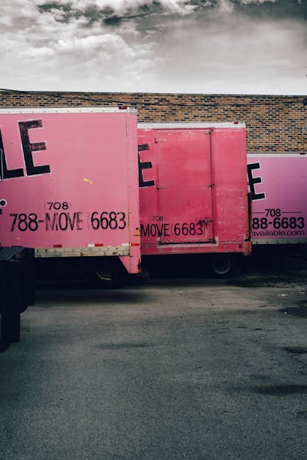 View of two large pink moving containers positioned on the pavement against a brick wall exterior of a property. The containers are made of sturdy materials with visible metal reinforcements and locking mechanisms, used for packing and transporting household items during a home relocation. One container is partially visible, overlapping another, with black text and contact details printed on their sides, including a phone number. The containers are part of the loading process, ready for transport. The background shows a cloudy sky, indicating outdoor conditions. The scene is associated with professional removals services offered by Man with Van Edgware, focusing on furniture transport and packing during house removals in the Edgware area.