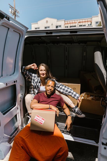 Two women are engaged in a home relocation process, seated at the rear of a large van with its back doors open. The woman in the foreground, with dark skin and wearing a red shirt, is holding a cardboard box labeled 'FRAGILE' and smiling at the camera while seated on an orange blanket. The woman behind her, with light skin, long brown hair, wearing a plaid shirt, is sitting inside the van, also smiling and looking out at the camera. The interior of the van is partially visible, showing several packed cardboard boxes and packing materials such as plastic wrap and paper. The setting appears to be outdoors in a residential area with a clear sky, and a multi-storey building is visible in the background. This scene illustrates the loading process of furniture and household belongings during house removals, aligned with services provided by Man with Van Edgware, supporting efficient and organized furniture transport and packing for home moves.