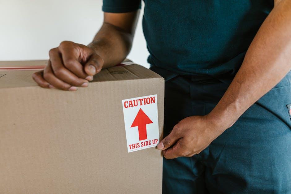 Close-up of a person wearing a dark T-shirt and blue trousers, holding the corner of a cardboard box during a home relocation. The box is sealed with packing tape and has a red and white caution label that reads 'Caution This Side Up' with an upward arrow. The individual is inside a room with a neutral background, likely preparing to load the box into a van or moving vehicle. The scene captures the careful handling and packing process involved in furniture transport and packing for a move, an essential part of same day removals services offered by Man with Van Edgware. The focus is on the proper orientation of packed items during the loading process, highlighting the importance of safety and organization during house removals.