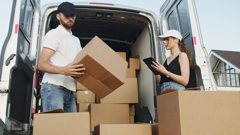 Two women are engaged in a home relocation process, seated at the rear of a large van with its back doors open. The woman in the foreground, with dark skin and wearing a red shirt, is holding a cardboard box labeled 'FRAGILE' and smiling at the camera while seated on an orange blanket. The woman behind her, with light skin, long brown hair, wearing a plaid shirt, is sitting inside the van, also smiling and looking out at the camera. The interior of the van is partially visible, showing several packed cardboard boxes and packing materials such as plastic wrap and paper. The setting appears to be outdoors in a residential area with a clear sky, and a multi-storey building is visible in the background. This scene illustrates the loading process of furniture and household belongings during house removals, aligned with services provided by Man with Van Edgware, supporting efficient and organized furniture transport and packing for home moves.